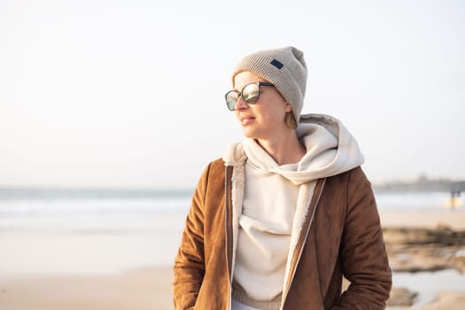Portrait of young stylish woman wearing brown padded jacket, hoodie, wool cap and sunglasses on long sandy beach in spring