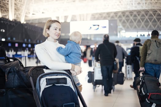 Motherat travelling with his infant baby boy child, walking, pushing baby stroller and luggage cart at airport terminal station. Travel with child concept