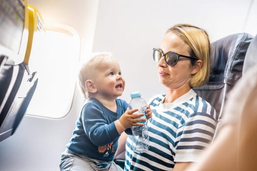 Mom and child flying by plane. Mother holding and playing with her infant baby boy child in her lap during economy comercial flight. Concept photo of air travel with baby. Real people