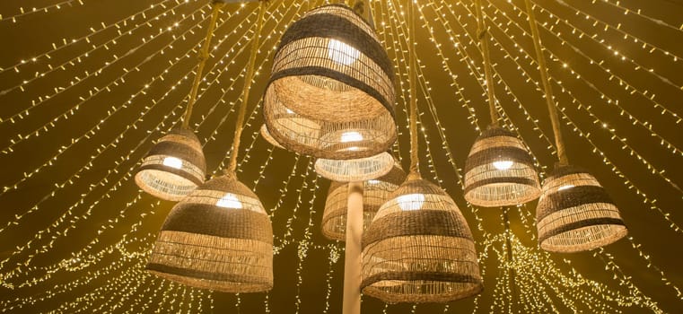 Natural fiber lamps hanging from the ceiling of a tent with a background of LED garlands. Warm atmosphere