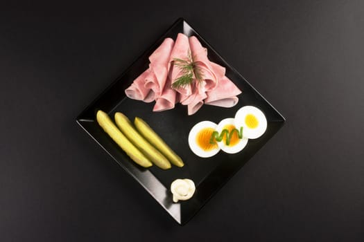 Elegant cold plate appetizer with ham, eggs and pickle on black plate on dark background.