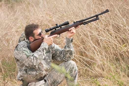 The hunter in military uniform is aiming and shooting a weapon with sniper in dry bush.