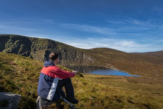 Men sitting on a rock and drinking coffee or tea from thermos. Scenic view on Lough Ouler and Tonelagee Mountain, hiking in Wicklow Mountains, Ireland