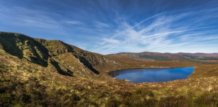 Wide panorama with rough hillside of Tonelagee Mountain and heart shaped lake, Lough Ouler, reflecting blue sky. Hiking in Wicklow Mountains, Ireland