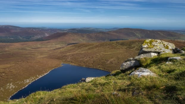 Stack of large rocks on a hillside of Tonelagee Mountain with a view on lake Lough Ouler, reflecting blue sky. Hiking in Wicklow Mountains, Ireland
