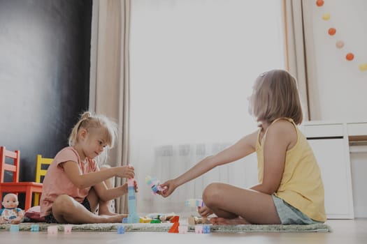 Children playing with colorful constructor building blocks, bricks. Kids sitting on floor indoors at sunny white home bedroom or kindergarten, play center. Funny educational child eco-friendly game