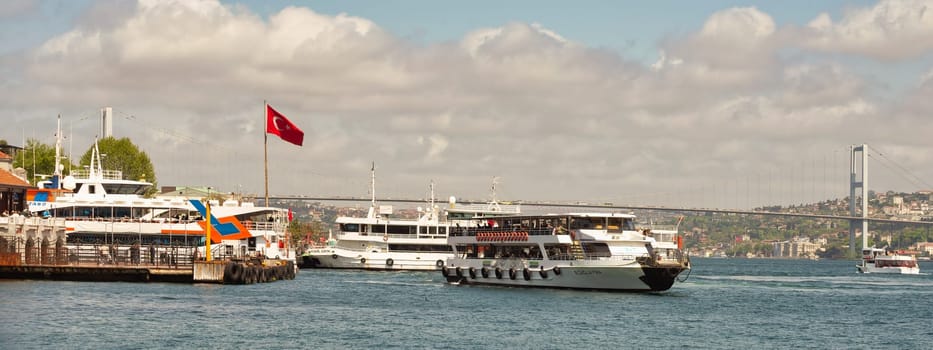 Istanbul, Turkey - May 7, 2023: Boats float peacefully on the Bosphorus Strait near Besiktas ferry terminal. The iconic Bosphorus Bridge towers in the background. The sky is cloudy in a spring day