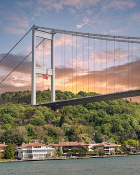 View from the sea of the Asian side of Bosphorus strait, Istanbul, Turkey, with traditional waterfront houses, under Bosphorus bridge, and green hill with dense trees before sunset