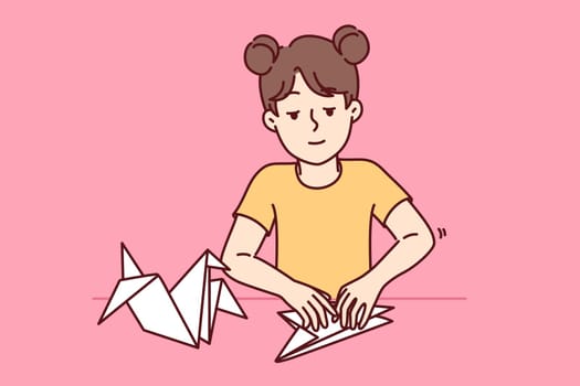 Little girl makes origami bird by folding paper animals and developing own creativity. Child is engaged in origami art, preparing fake for school exhibition and dreaming of becoming sculptor