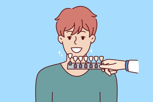 Man at dentist office showing teeth for comparison with dental palette in preparation for whitening procedure. Young guy visits dentist wanting to improve smile or get rid of toothache.