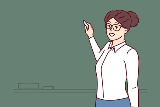 Woman teacher stands at blackboard and uses chalk, teaching elementary or high school children and giving students new knowledge. Happy teacher posing near empty chalkboard before class starts