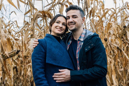 A happy couple in love stands in autumn near a cornfield. A man in a shirt and jacket hugs a woman in a coat.
