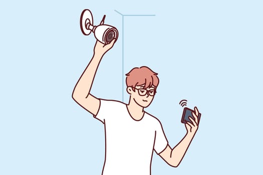 Man installs surveillance camera on wall and holds mobile phone, watching video through WIFI network. Guy establishes video surveillance system, wanting to secure home or office from criminals.