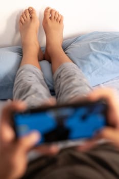 A teenage boy in mid-morning pajamas lying on the bed is playing video games with his feet resting on the pillow. Focus on background and copy space.