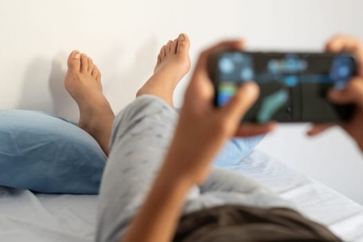 Barefoot teenage boy with crossed legs resting on pillow using phone lying on bed. Teenaged male playing video games in his room mid-morning.