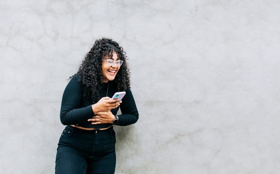 Pretty girl happy with her cell phone, young latin woman happy with her cell phone leaning against a wall. A young latin woman laughing with her cell phone leaning against a wall