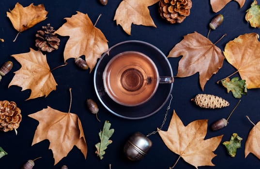 Cup of tea with autumn leaves, acorn and cones on dark background from above. Maple leaf texture on black paper. Seasonal composition pattern design with cozy drink. Flat lay.