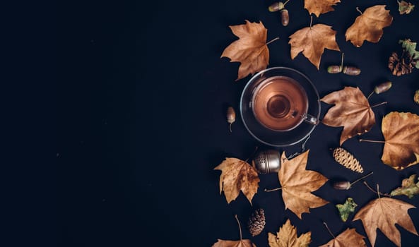 Cup of tea with autumn leaves, acorn and cones on dark background from above. Maple leaf texture on black paper. Seasonal composition pattern border design with cozy drink. Flat lay.