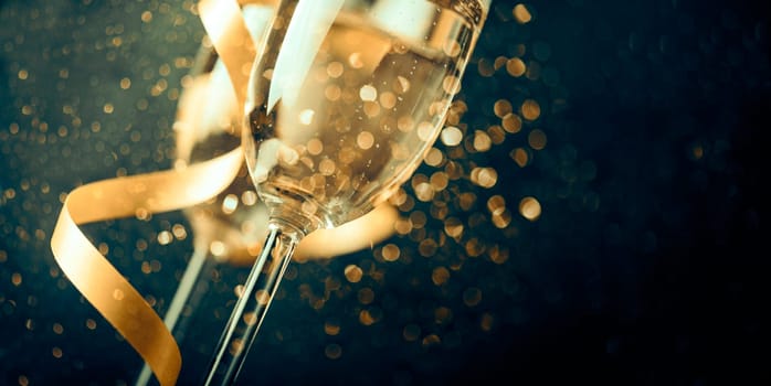 Two champagne glasses with golden ribbon and gold glitter splash bokeh on dark background. Luxury restaurant dinner celebration. Classy christmas and new year holiday panoramic design banner.