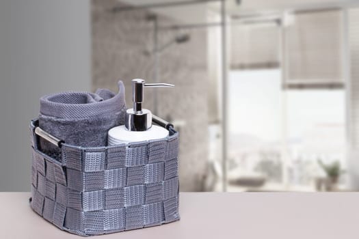 Basket with a fresh soft terry bath towel and a soap bottle on a white table over blurred bath background with copy space. For your product display montage.