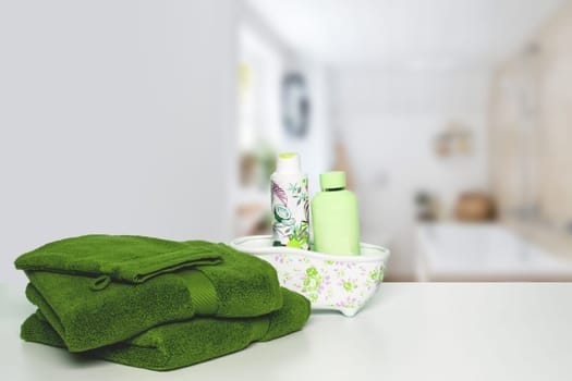 A stack or pile of green soft terry bath towels and beauty cosmetic makeup bottles on bright table  over blurred bathroom background space. Beauty and Wellness concept.