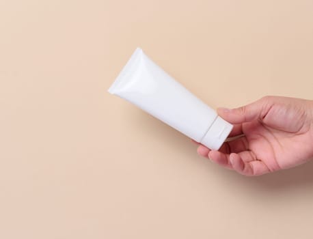 A woman's hand holds a white plastic tube for cream, toothpaste on a beige background