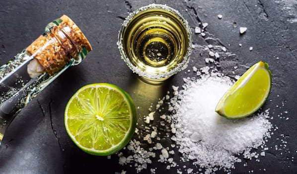 Mexican Tequila Tres Sombreros bottle with lime, salt and shot glasses closeup against dark background
