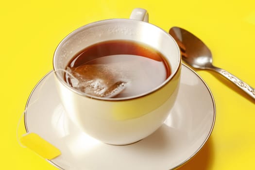 White porcelain cup with hot freshly brewed tea, bag still in, silver spoon next on yellow board, lit by morning sun