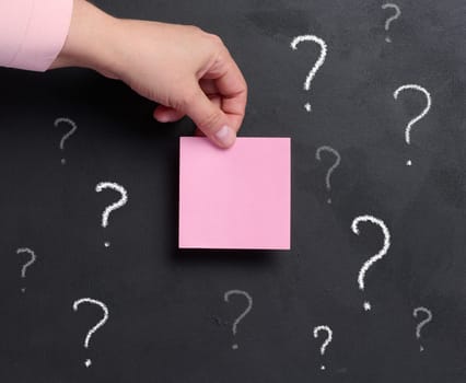 A woman's hand holds a pink paper sticker on a background of a black chalk board with drawn question marks.