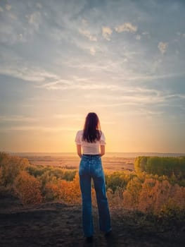 Rear view of a young woman on te top of a hill admiring the calm sunset over the autumn valley with colorful trees. Fall season melancholy feeling, copy space sky background