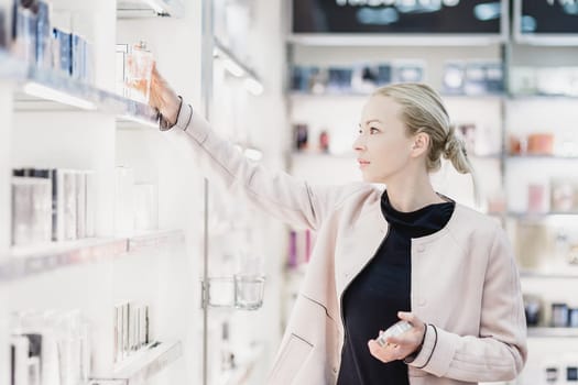 Elegant blond young woman choosing perfume in retail store. Beautiful blond lady testing and buying cosmetics in a beauty store.