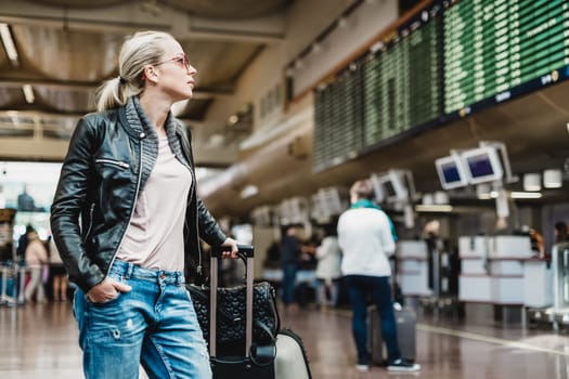 Casually dressed young stylish female traveller checking a departures board at the airport terminal hall in front of check in couters. Flight schedule display blured in the background. Focus on woman.