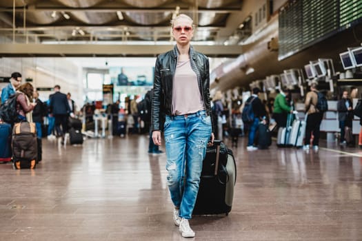 Casually dressed young stylish female traveler walking the airport terminal hall dragging suitcase and a handbag behind her. Blurred background. Can also be used as railway, metro, bus station.