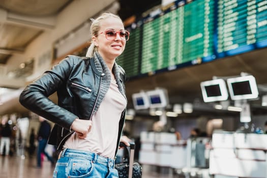 Casually dressed young stylish female traveller checking a departures board at the airport terminal hall in front of check in couters. Flight schedule display blured in the background. Focus on woman.