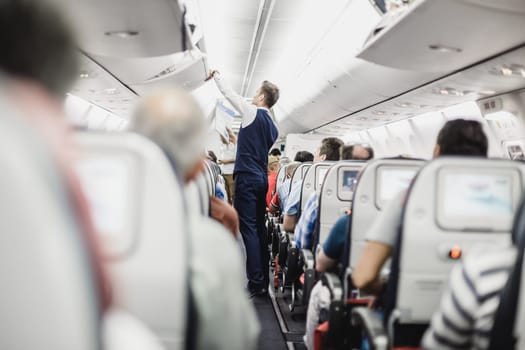 Interior of airplane with passengers on seats and steward in uniform closing overhead luggage compartment.