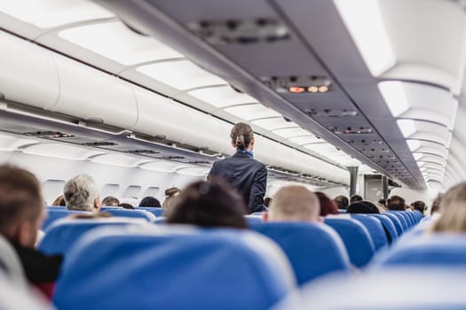 Interior of airplane with passengers on seats and stewardess walking the aisle.