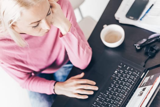 Female freelancer in her casual home clothing remotly working on laptop computer from her home while drinking her morning cup coffee. View from above.