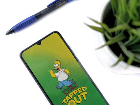 Lod, Israel - July 16,2023: Modern workspace with smartphone with The Simpsons Tapped Out app launch screen on white background. Close up top view flat lay.