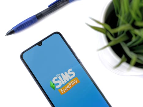 Lod, Israel - July 16,2023: Modern workspace with smartphone with The Sims FreePlay app launch screen on white background. Close up top view flat lay.