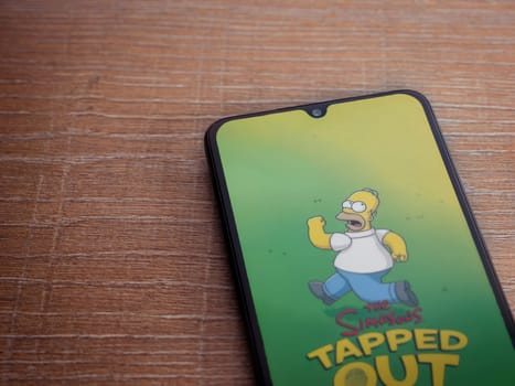 Lod, Israel - July 16,2023: The Simpsons Tapped Out app launch screen on smartphone on wooden background. Top view flat lay with copy space.