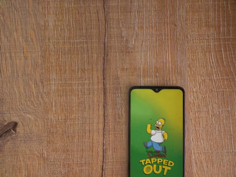 Lod, Israel - July 16,2023: The Simpsons Tapped Out app launch screen on smartphone on wooden background. Top view flat lay with copy space.
