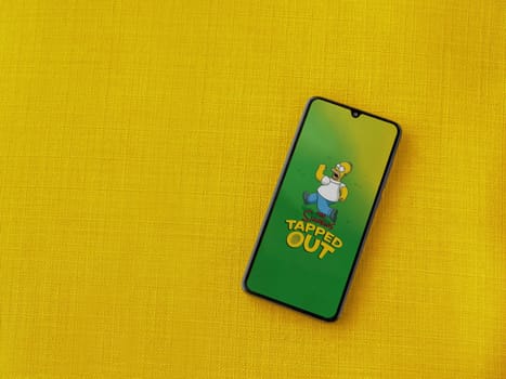 Lod, Israel - July 16,2023: The Simpsons Tapped Out app launch screen on smartphone on yellow fabric background. Top view flat lay with copy space.