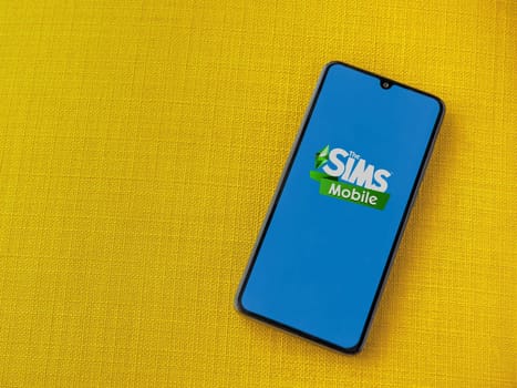 Lod, Israel - July 16,2023: The Sims Mobile app launch screen on smartphone on yellow fabric background. Top view flat lay with copy space.
