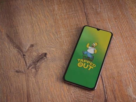 Lod, Israel - July 16,2023: The Simpsons Tapped Out app launch screen on smartphone on wooden background. Top view flat lay with copy space.
