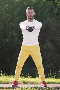 Front view of the sporty young guy is exercising with kettlebell sport equipment outdoors.