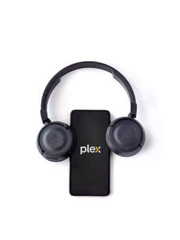 Lod, Israel - July 16,2023: Smartphone with Plex app launch screen and wireless headphones on white background. Top view flat lay with copy space.