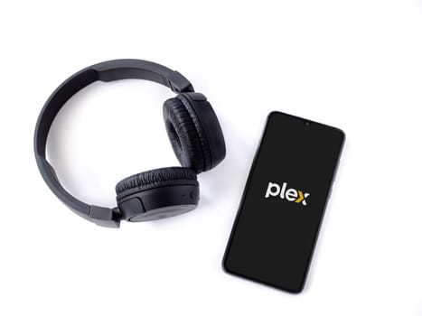 Lod, Israel - July 16,2023: Smartphone with Plex app launch screen and wireless headphones on white background. Top view flat lay with copy space.