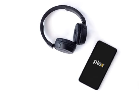 Lod, Israel - July 16,2023: Smartphone with Plex app launch screen and wireless headphones on white background. Top view flat lay with copy space.