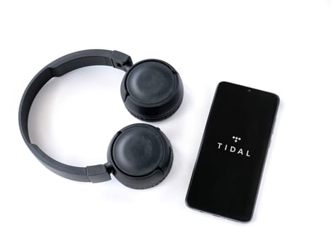 Lod, Israel - July 16,2023: Smartphone with TIDAL Music app launch screen and wireless headphones on white background. Top view flat lay with copy space.