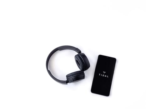 Lod, Israel - July 16,2023: Smartphone with TIDAL Music app launch screen and wireless headphones on white background. Top view flat lay with copy space.